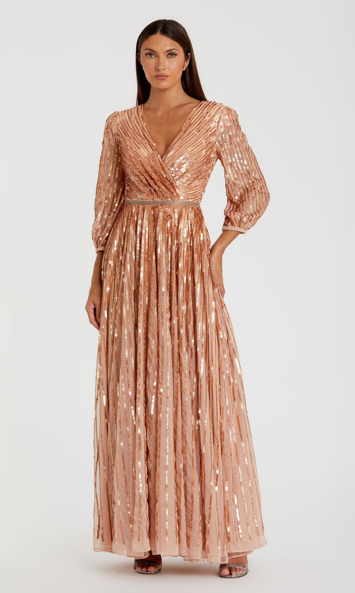 A woman stands against a plain background wearing the Mac Duggal 5509, a long sleeve formal dress with a V-neck, floor-length rose gold sequins, and a pleated skirt, paired with open-toe heels.